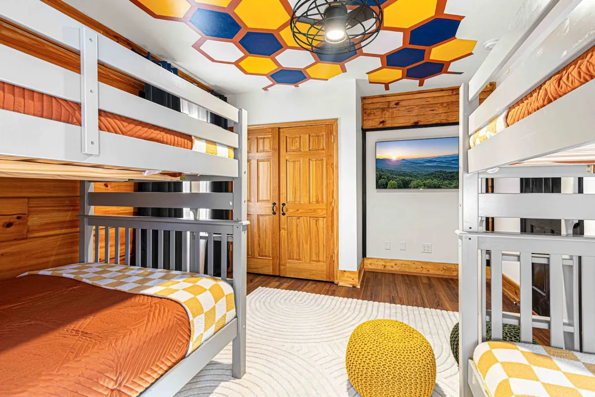 A playful and cozy loft-style room with colorful bunk beds, a TV displaying landscape artwork, a textured rug, and decorative poufs. The space is inviting for both sleep and activities such as storytime or movie nights.