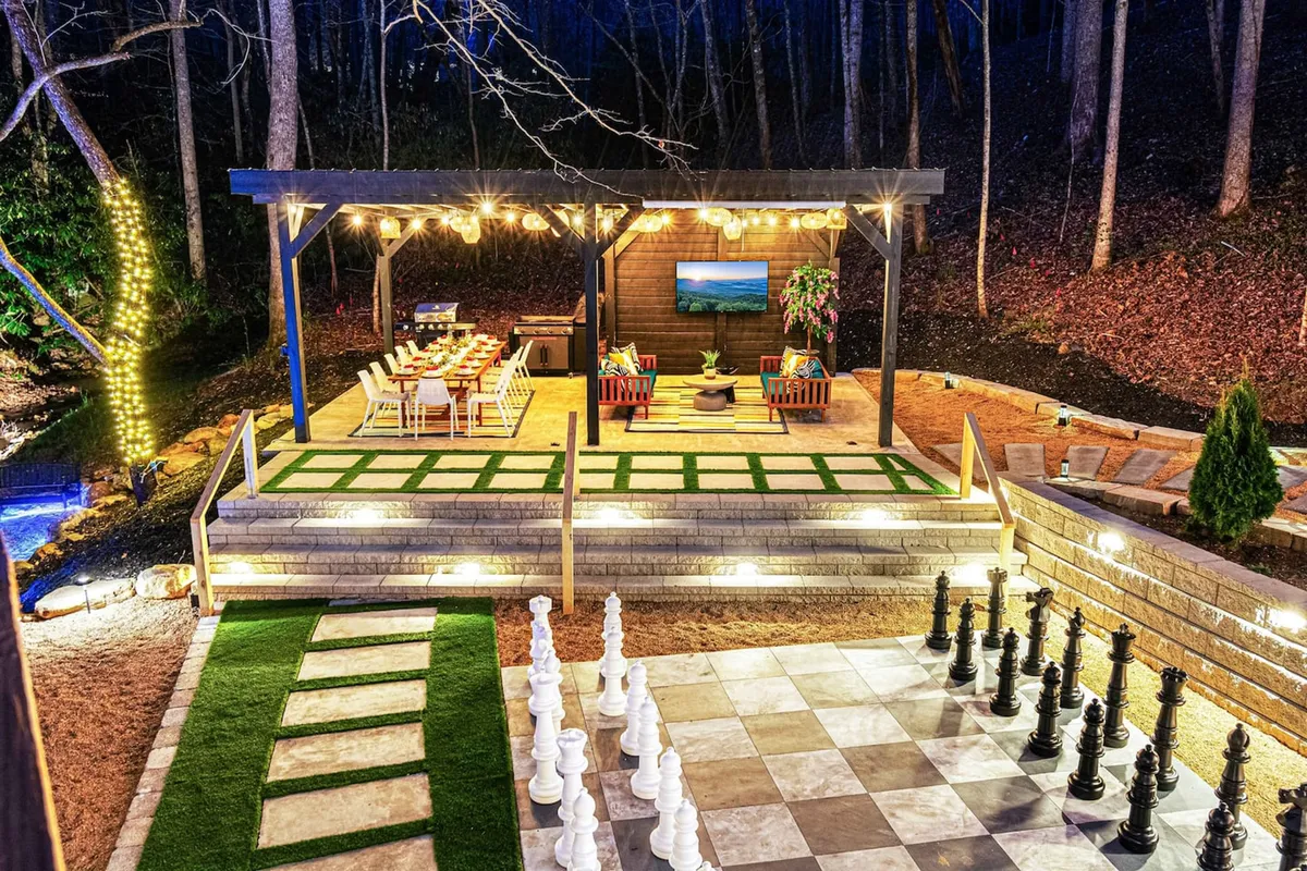 A scenic outdoor setting featuring a dining area under a pergola, a chess set for leisure, decorative string lights, and comfortable seating. The space is designed for relaxed socializing and festive al fresco dining amid a refined atmosphere.