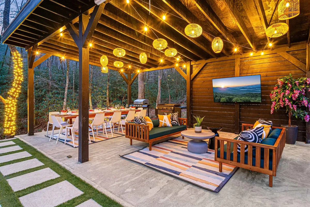 An inviting exterior setup with hanging pendant lights, an outdoor dining table, comfortable seating, and a large television, all arranged to create a warm, festive atmosphere. The scene is softly illuminated for sunset relaxation or evening gatherings, offering a seamless blend of nature and luxury.