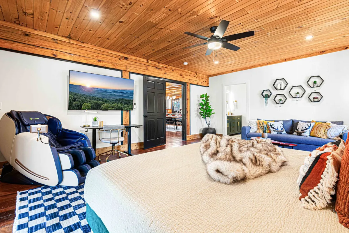 A serene bedroom featuring a king-sized bed draped with a textured blanket, a plush blue sofa, a large flat-screen TV displaying scenic views, a luxurious massage chair, and decorative wall shelves. The room radiates comfort, blending relaxation and entertainment spaces seamlessly.
