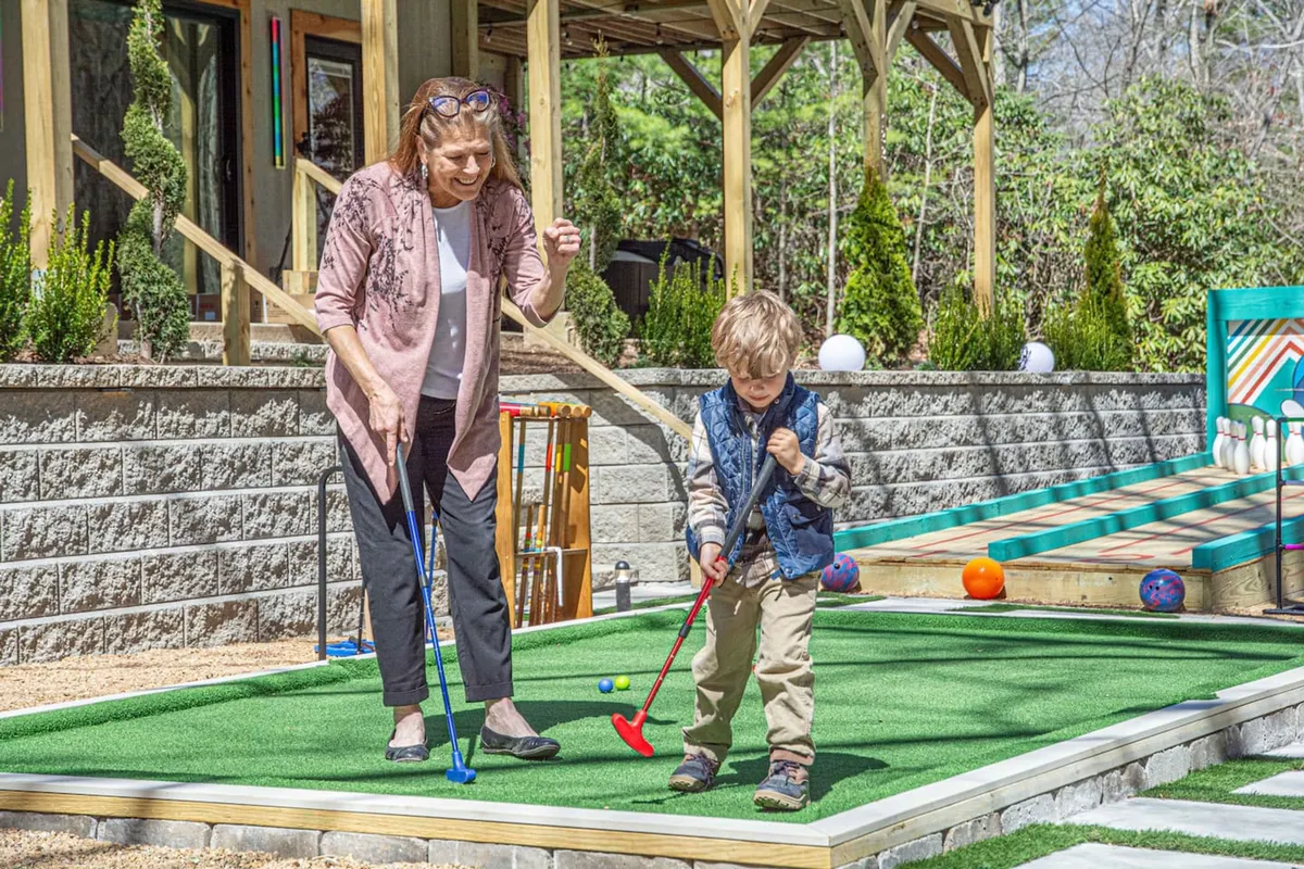 A joyful scene of a grandmother and child playing mini-golf on a green artificial turf, with a colorful outdoor bowling setup in the background. The setting is bright and playful, emphasizing multi-generational family fun and outdoor activities.