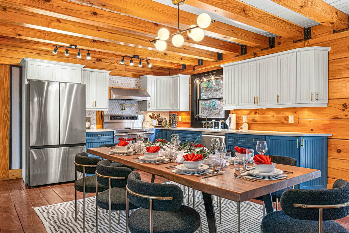 The scene shows a dining table beautifully set for a meal, adjacent to a kitchen featuring modern appliances and wooden beams. The space exudes warmth and hospitality, with ample room for friends and family to gather, cook, and dine together.