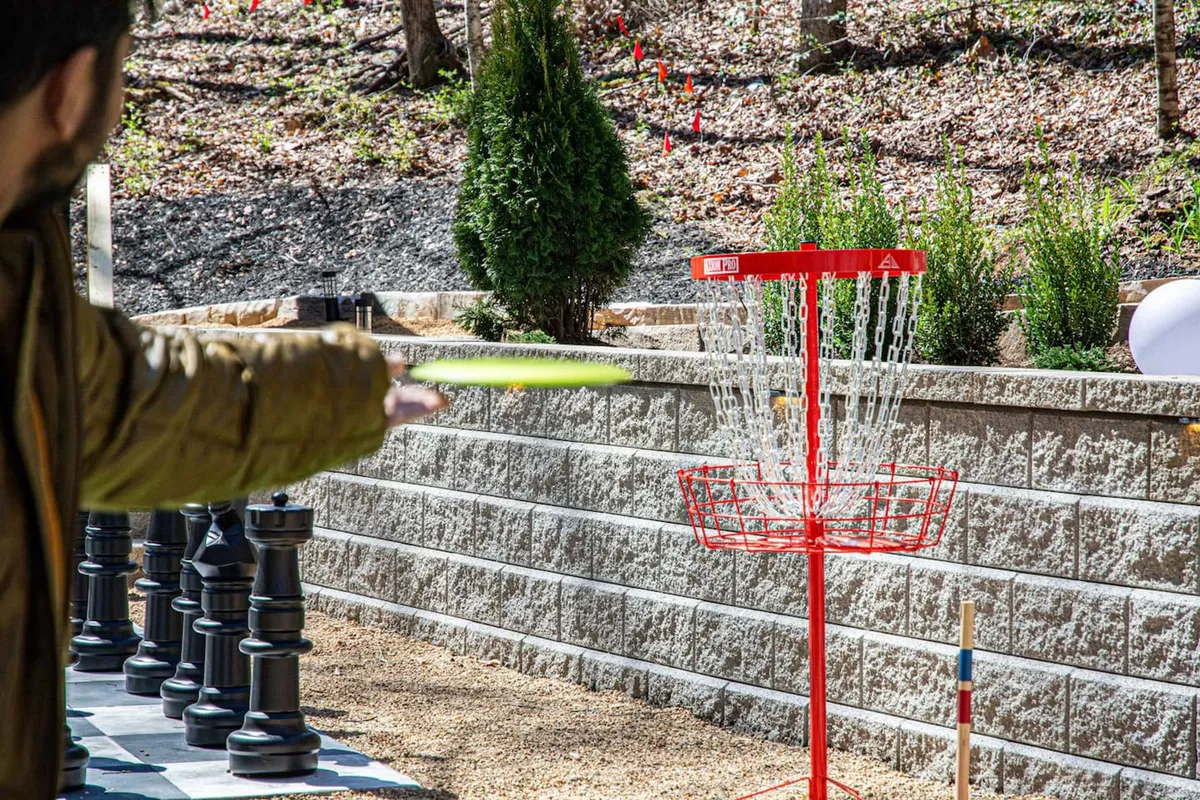An open-air recreational area featuring a disc golf basket, a person mid-throw, and oversize chess pieces, all set in a spacious, green environment. The scene evokes a sense of playful exploration and friendly competition outdoors.