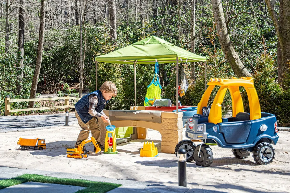 A close-up of a child playing joyfully in a sandbox, surrounded by a play car and shaded by a green canopy. The sandbox area is inviting, colorful, and clearly designed for creative, hands-on play, making it an ideal spot for younger kids to dig, build, and explore.