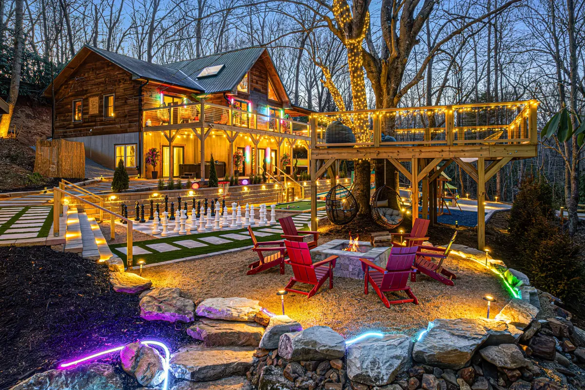 The image depicts an illuminated outdoor deck at dusk, featuring a large chess set, a cozy fire pit surrounded by Adirondack chairs, and hanging chairs set for relaxation. The atmosphere is playful and welcoming, perfect for evening gatherings or group games under the stars.