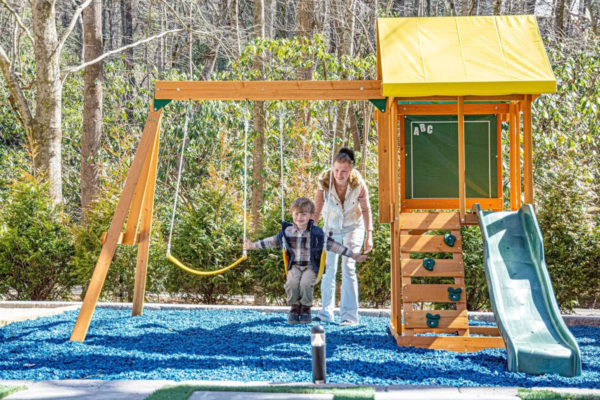 A vibrant outdoor playground scene featuring a play structure with swings and a slide, children actively playing, and a parent nearby. The space is surrounded by greenery, emphasizing a safe and welcoming environment for children and families to gather and create memories together.