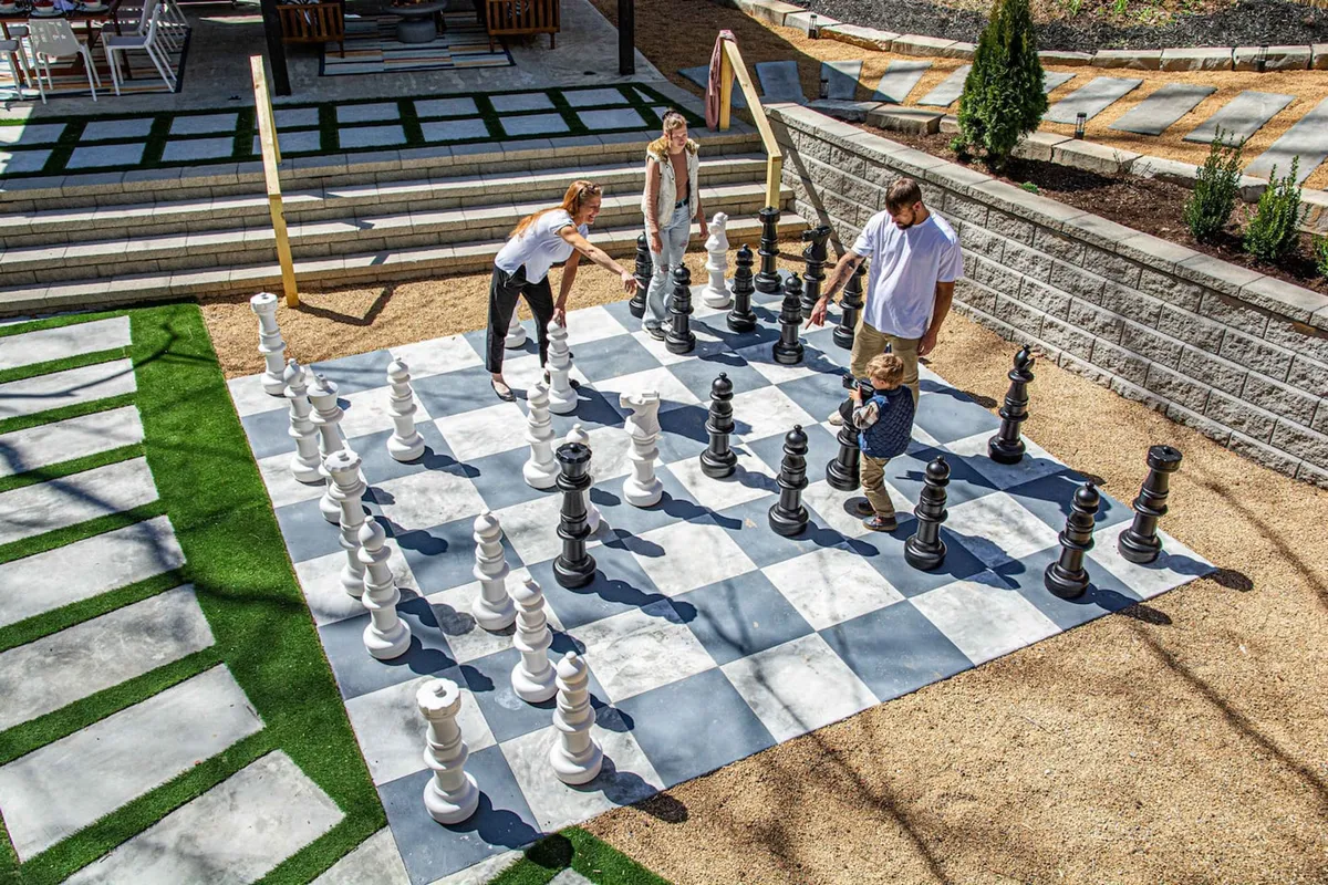 An exterior shot of a large outdoor chess set where family members are engaged in play on a green lawn surrounded by stone pathways. The mood is lively and interactive, highlighting outdoor leisure and learning moments between parents and children.