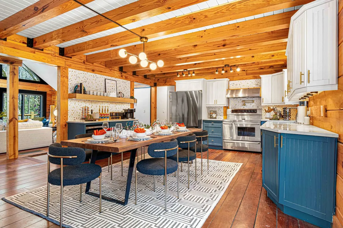 This image captures a dining table positioned beside a kitchen area equipped with modern appliances and wooden beams overhead. Decorative backsplash and warm lighting add to the inviting atmosphere, with the table set up for dining and ready for gatherings.