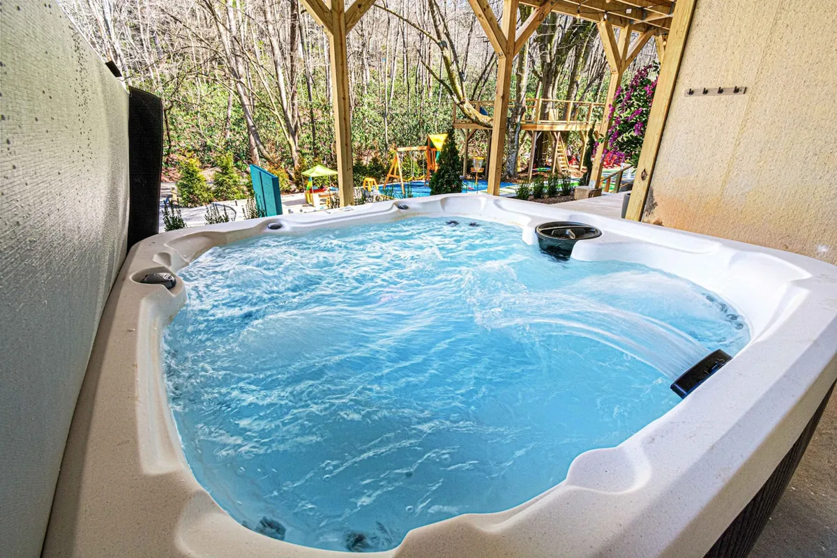 An inviting hot tub on a deck is set against a backdrop of natural greenery, with playground equipment visible nearby. The scene captures the dual appeal of relaxation for adults and play for children, suggesting the versatility of the yard as a space for all ages to enjoy together.